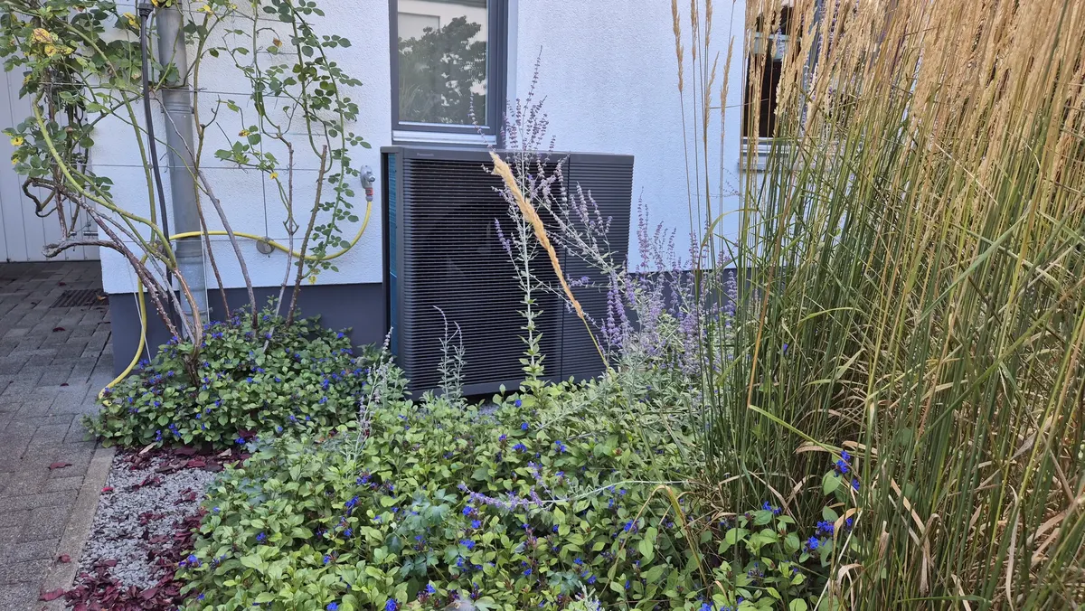 Dunkle Panasonic Wärmepumpe im bepflanzten Garten vor dem Haus